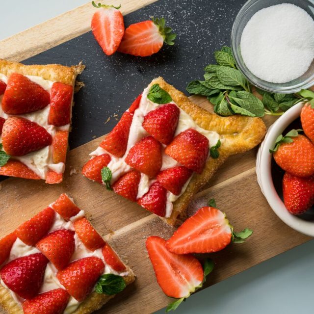 Three slices of strawberries and cream puff pastry on a wood board with fresh strawberries, mint and sugar