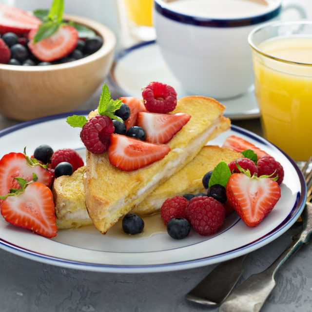 Baked stuffed French toast with cream cheese filling topped with strawberries, raspberries and blueberries beside orange juice and coffee