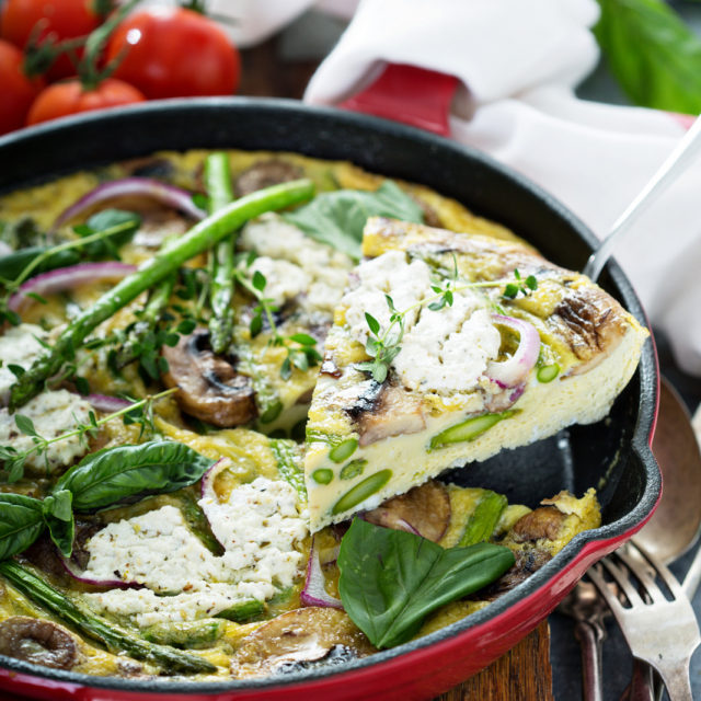 Asparagus, goat cheese and mushroom frittata in a red skillet with a slice being lifted out