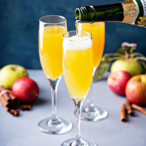 Sparkling wine being poured from a green bottle into a flute of apple cider, with red and green apples and cinnamon sticks on the table