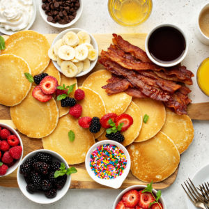 Overhead pancake board with bacon, syrup, berries, sprinkles and forks
