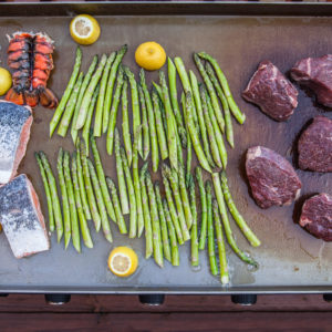 Raw filet mignon steaks, lobster tails, salmon fillets, asparagus, and lemons arranged on the griddle