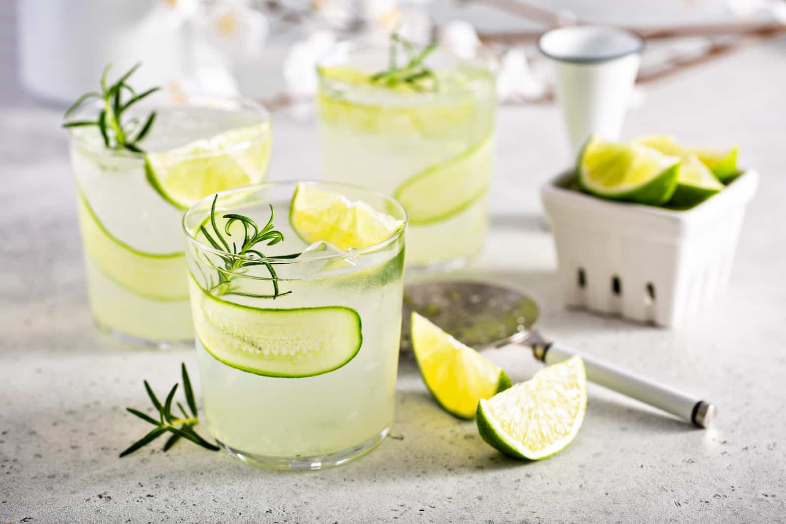 Three cucumber gin gimlet cocktails with cucumber ribbons, lime wedges and rosemary in rocks glasses