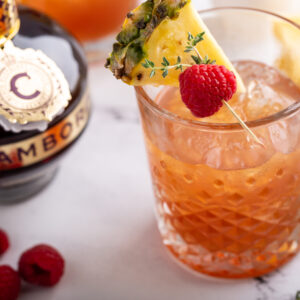 Chambord bottle beside a sweet thyme cocktail with pineapple and raspberry