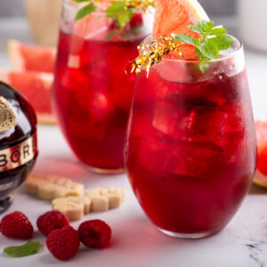 Two raspberry bourbon sangrias in stemless glasses with grapefruit and mint beside a Chambord bottle and fresh raspberries