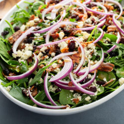 Overhead of a spinach and arugula salad with red onion rings, dried cranberries, pecans, bacon and Gorgonzola in a white oval bowl beside wooden servers