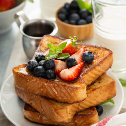Stack of golden French toast topped with fresh strawberries, blueberries and mint on a white plate