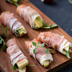 Close-up of prosciutto-wrapped apple bites on a walnut board with red holiday ornaments behind them