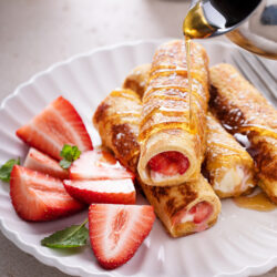 Syrup being poured over strawberry French toast roll-ups with sliced strawberries