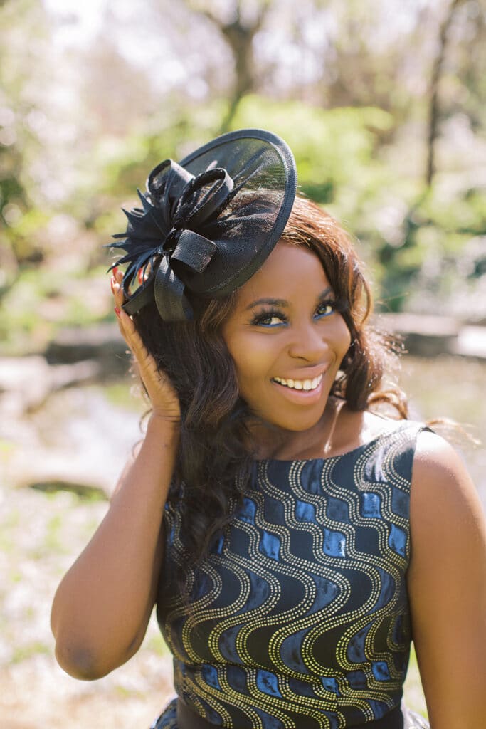 Erica Thomas touching the brim of her black fascinator, smiling in a spring garden
