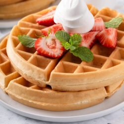 Close-up of a stack of waffles topped with whipped cream, sliced strawberries and fresh mint