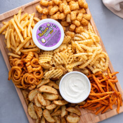 Overhead French fry board on a wooden cutting board with seven kinds of fries, a tub of Heluva Good ranch dip and a bowl of dip, beside a white linen towel