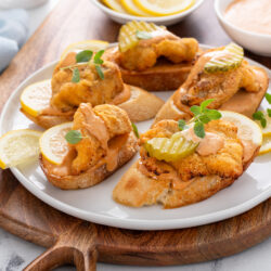Plate of fried catfish nugget crostini topped with remoulade sauce and pickle slices, garnished with lemon and fresh oregano