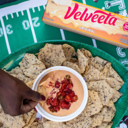 Bowl of spicy queso dip topped with diced tomatoes, surrounded by tortilla chips, with a box of Velveeta Original on a football-field backdrop