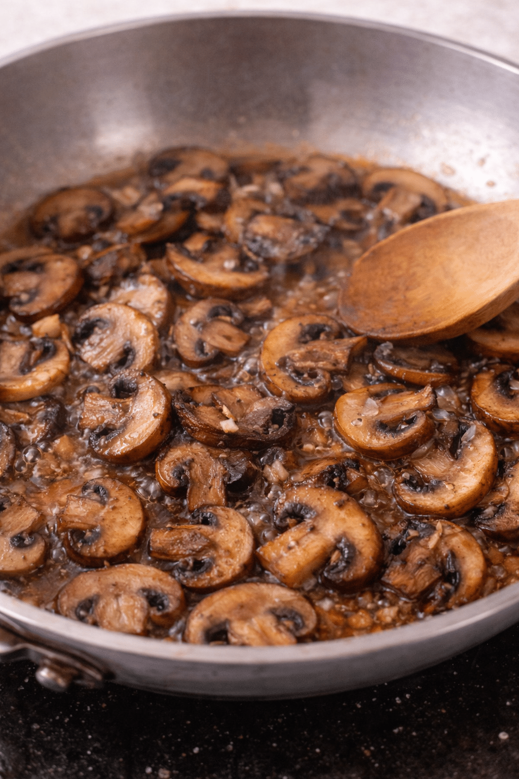 Sliced mushrooms browning in a skillet with a wooden spoon