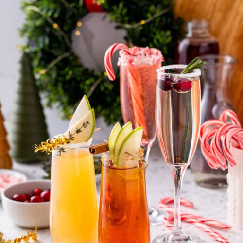 Three holiday mimosas on a speckled counter, garnished with pear slices, cranberries and rosemary, surrounded by candy canes and ornaments with a boxwood wreath behind