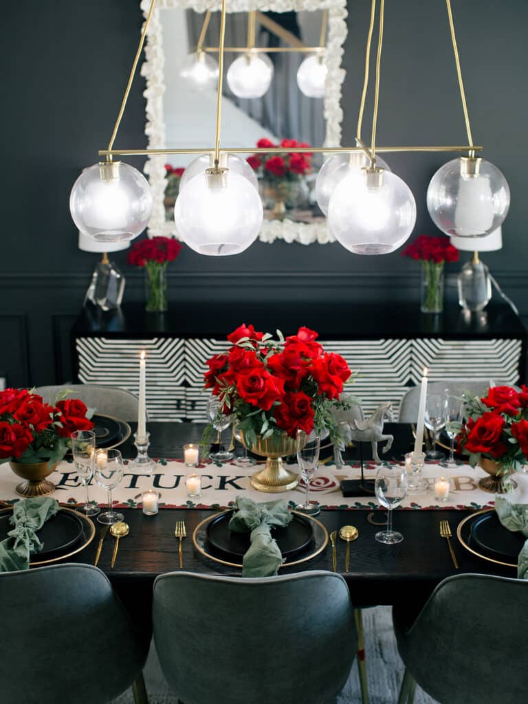 Kentucky Derby table seen head-on, with three red rose centerpieces along a Kentucky Derby runner and black and gold place settings