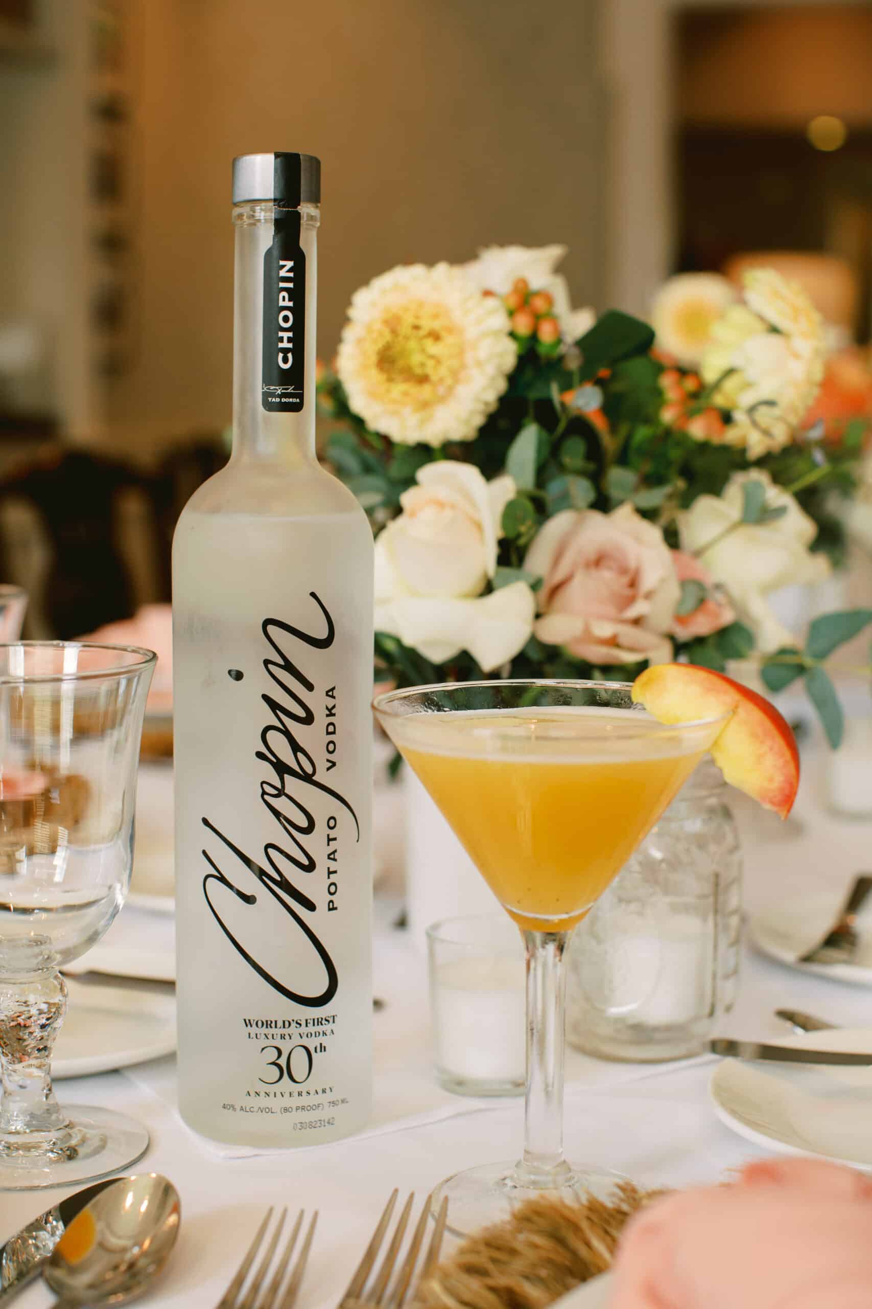 Peach martini garnished with a peach slice beside a bottle of Chopin vodka and peach-toned flowers on a set table
