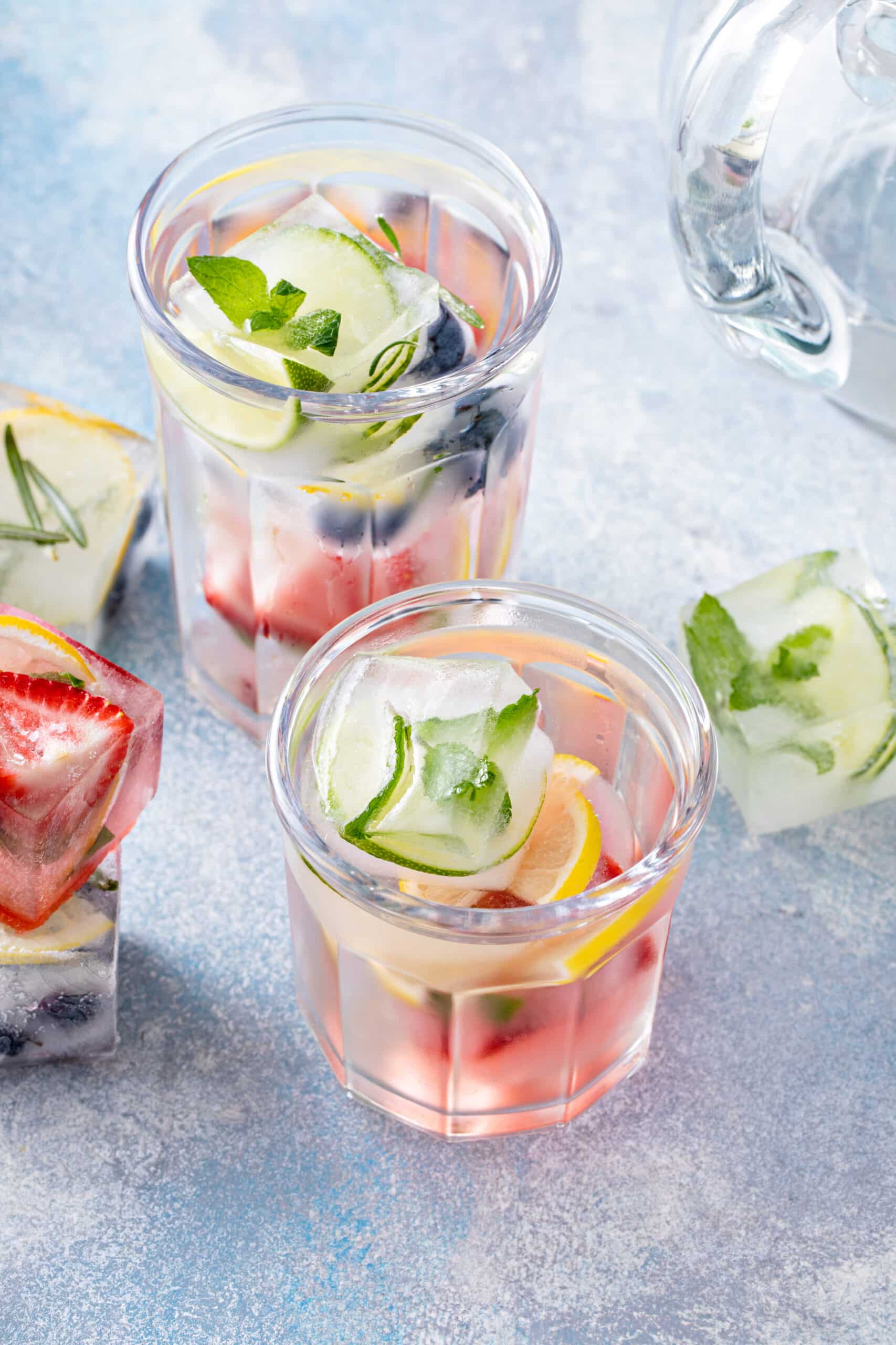 Two glasses of water with colorful fruit-and-herb ice cubes beside a pitcher