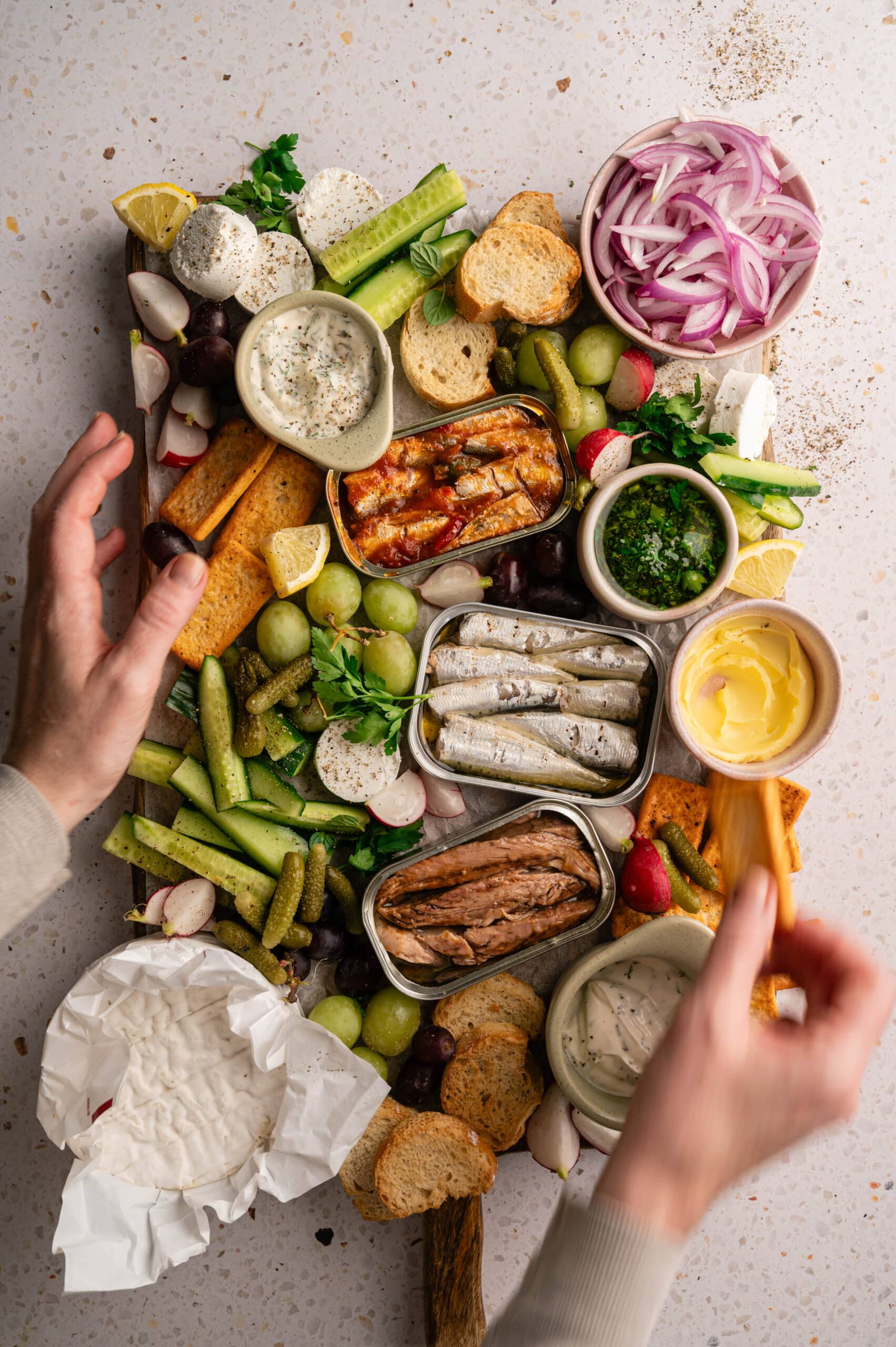 Hand reaching for a cracker on a tinned fish board with sardines, olives, cucumber and dips