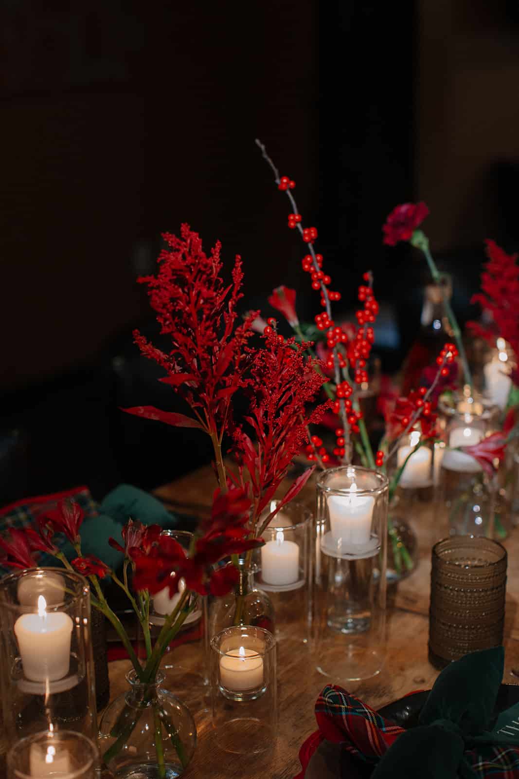 A dark holiday dinner table glowing with clusters of glass votive candles, red berry branches and red flowers in bud vases, and tartan napkins