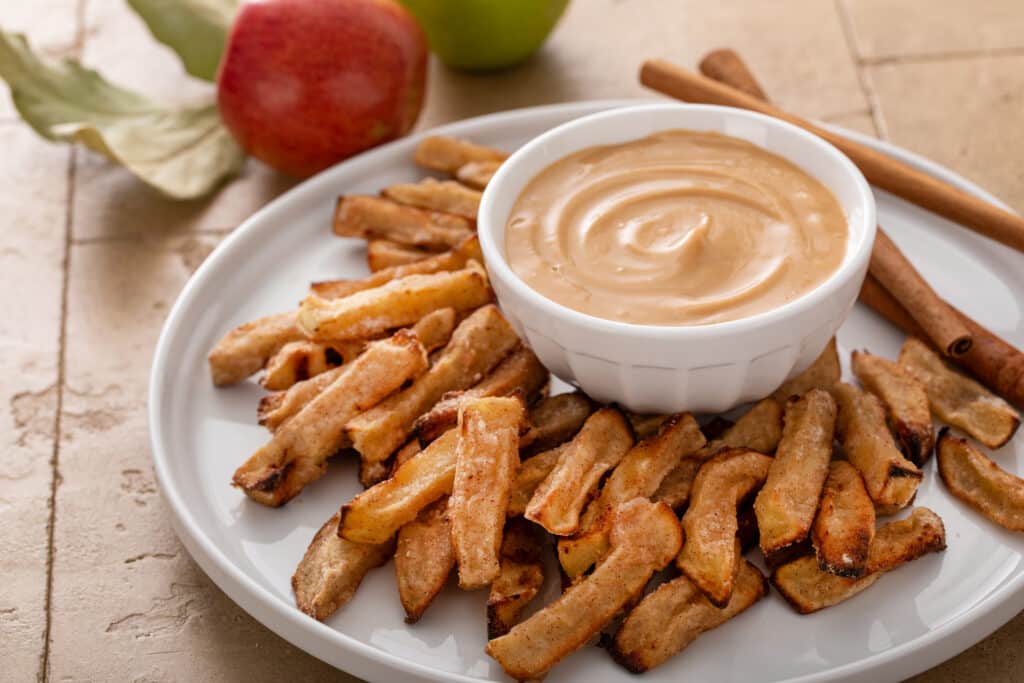 Air Fryer Apple Fries with Caramel Cream Dip on white serving plate with cinnamon sticks and red and green apple