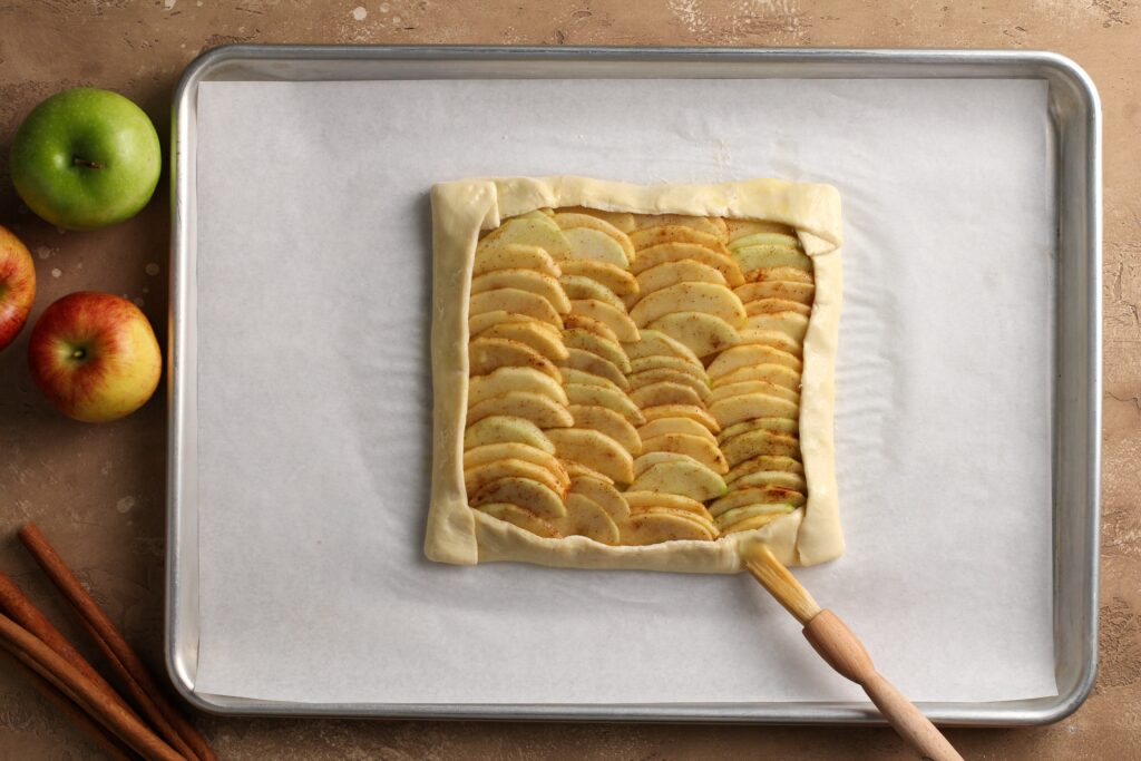 Unbaked apple galette being brushed with egg wash on a parchment-lined sheet pan with apples and cinnamon sticks