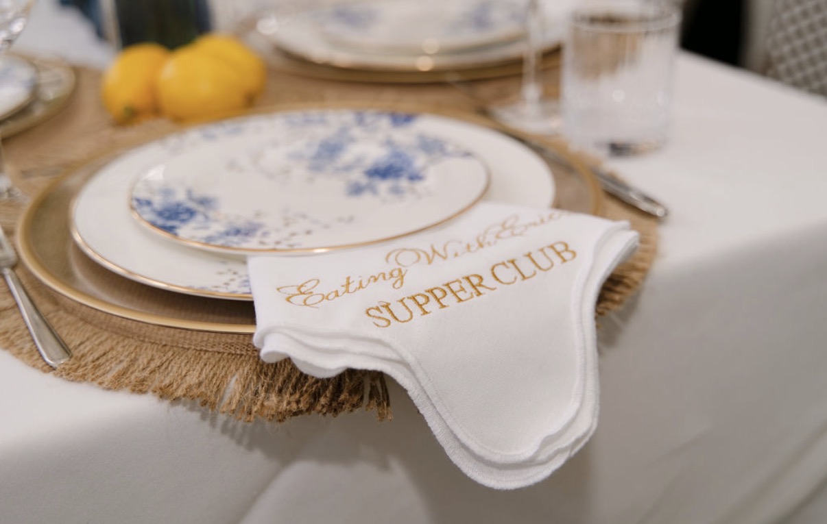 Close-up of a scalloped white napkin embroidered in gold with the Eating With Erica Supper Club logo on Lenox Garden Grove china