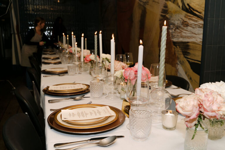 Long Supper Club table at Foundation Social Eatery set with gold-rimmed plates, printed menus, white and sage twisted taper candles and pink roses