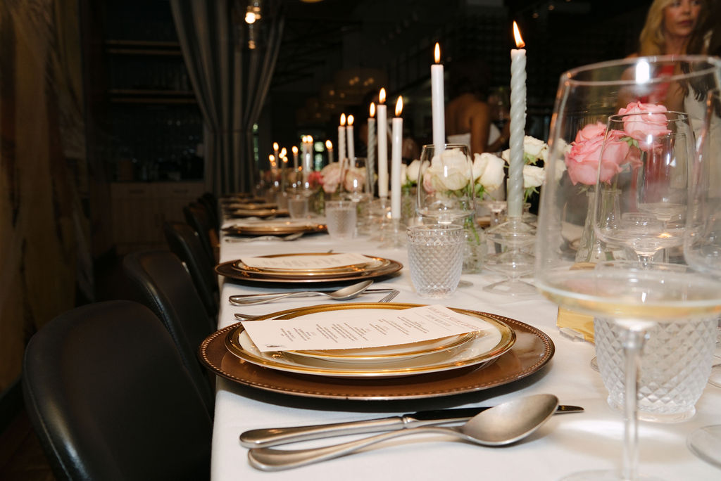 Row of place settings with printed menus, crystal tumblers, taper candles and pink and white roses at the Supper Club dinner