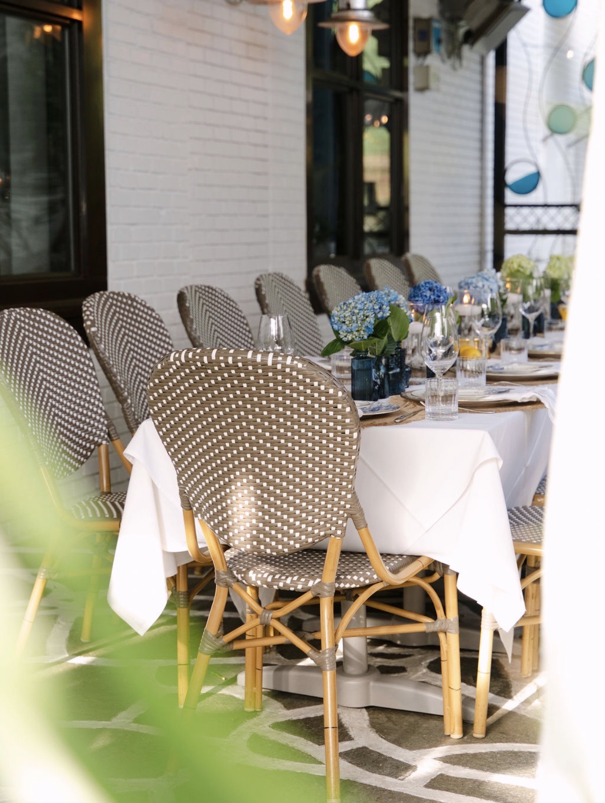 Woven bistro chairs at a white-linen dinner table with blue hydrangeas on a stone patio