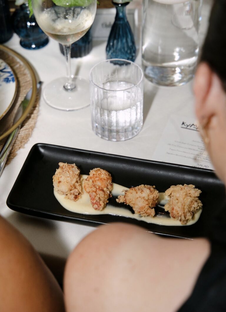 A plate of Kyma's crispy lobster morsels on the Supper Club table beside a Kyma menu, a glass of white wine and blue bud vases