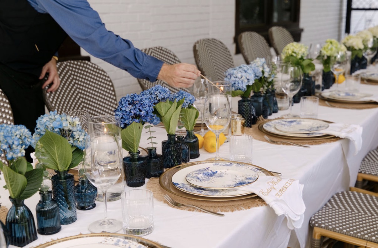 A Kyma server lighting floating candles along the Eating With Erica Supper Club table in Buckhead, set with blue hydrangeas, lemons and Lenox Garden Grove china