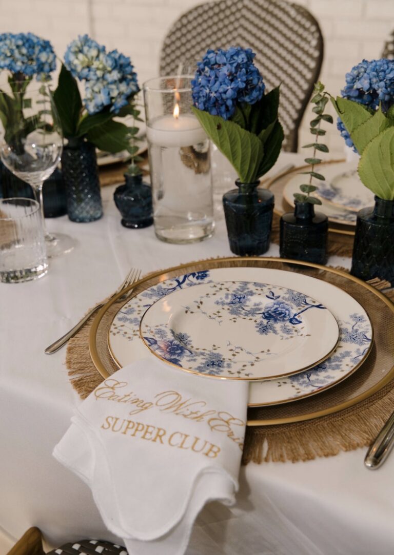 Lenox Garden Grove blue floral china with gold rims on a woven charger, with a white napkin embroidered in gold with the Eating With Erica Supper Club logo and blue hydrangeas behind