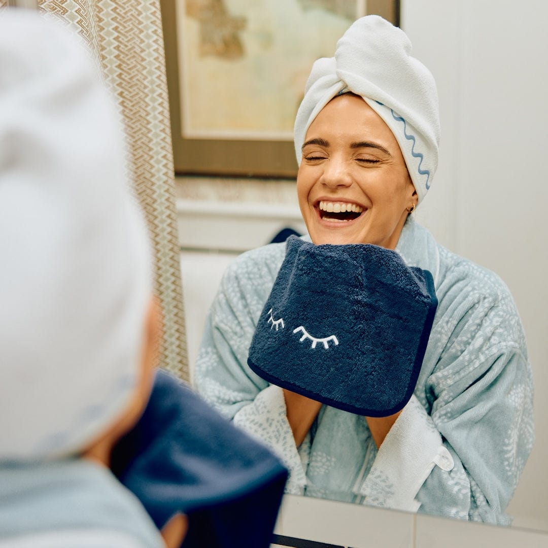 Woman with her hair wrapped in a white towel holding a navy Weezie makeup towel embroidered with closed eyes