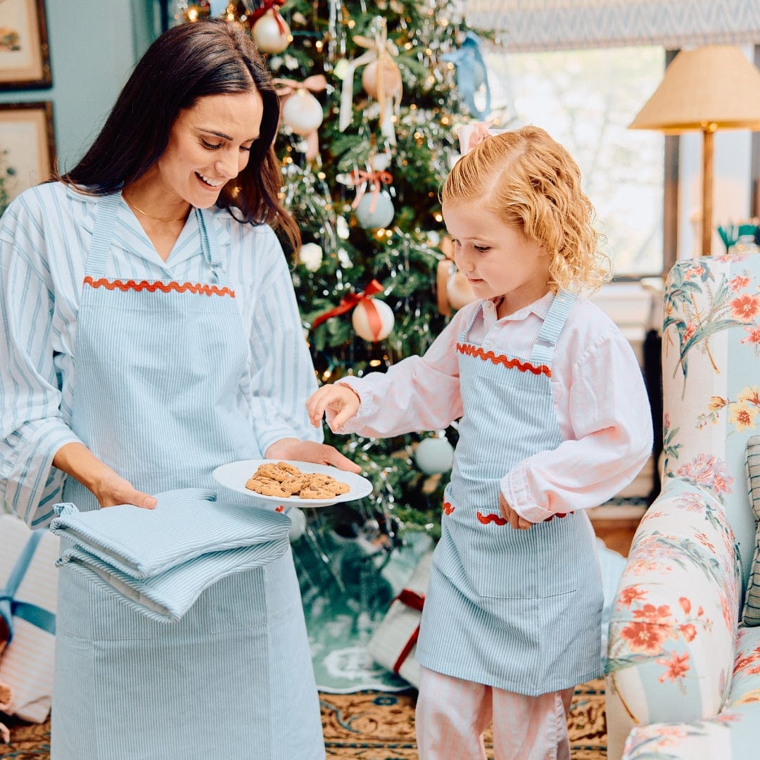 Mother and young daughter in matching light blue Weezie aprons with red trim holding a plate of cookies by a Christmas tree