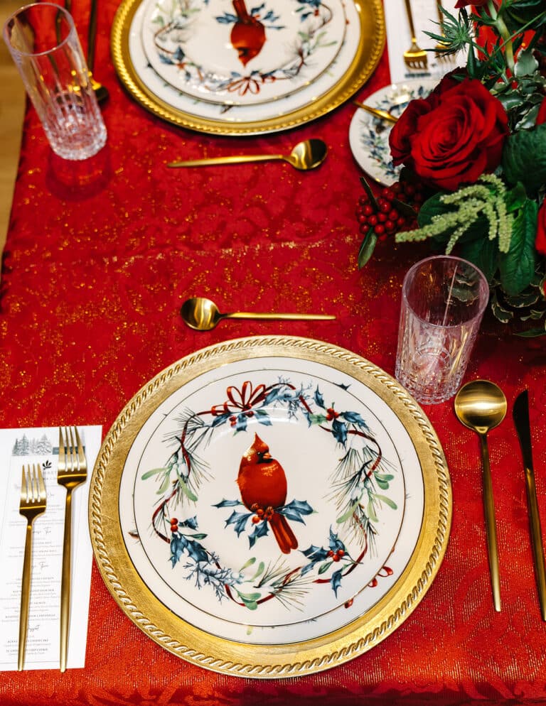 Close-up of a Lenox Winter Greetings cardinal plate on a gold charger with gold flatware, a printed menu and crystal glassware on a red holiday tablecloth