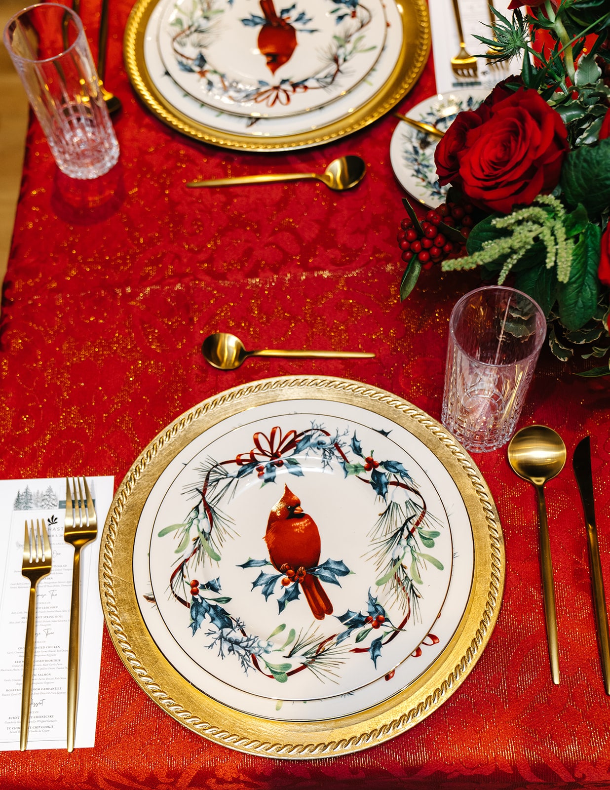 Close-up of a Lenox Winter Greetings cardinal plate on a gold charger with gold flatware, a printed menu and crystal glassware on a red holiday tablecloth