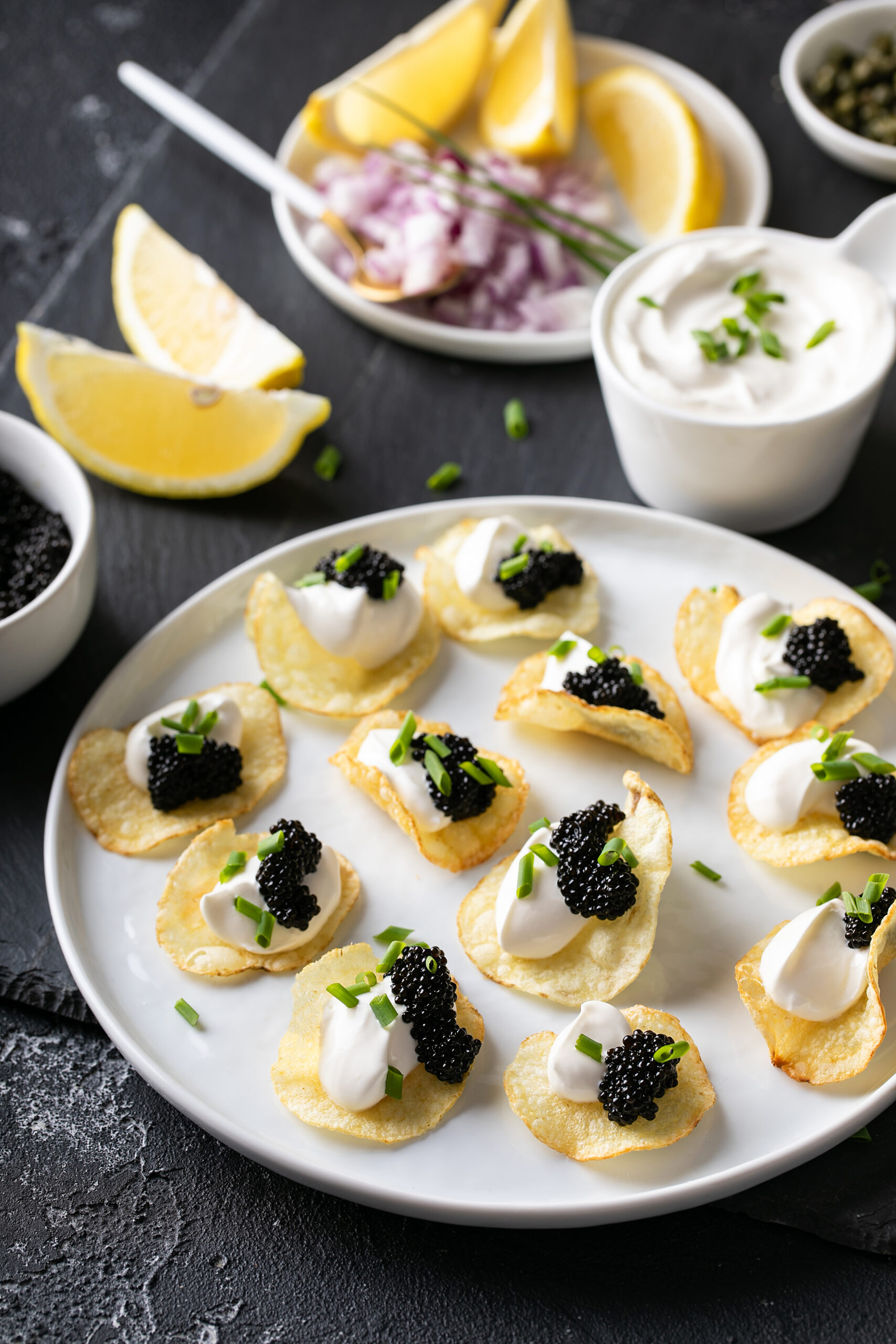 Plate of caviar potato chips with lemon wedges, diced red onion and a small pitcher of crème fraîche