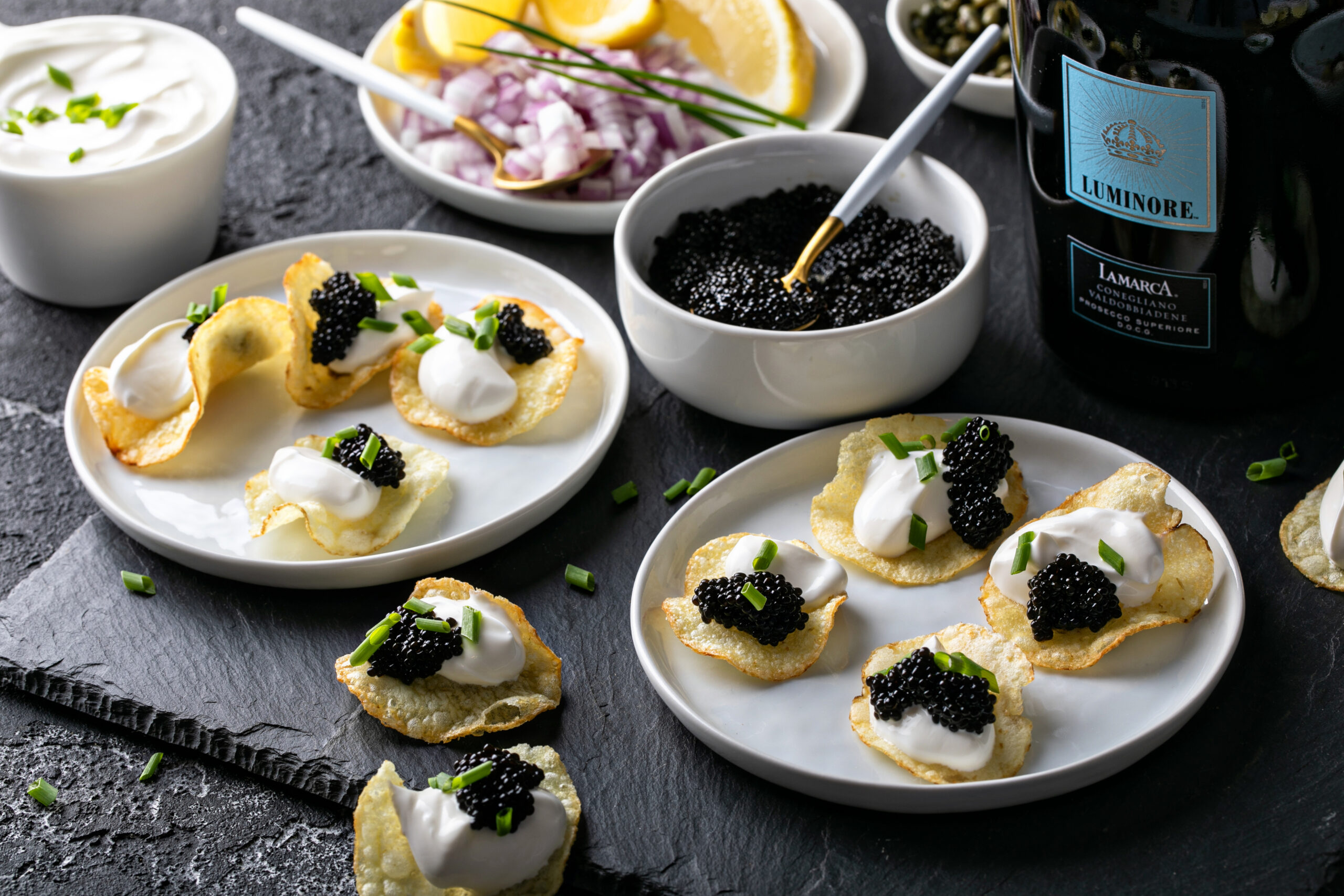 Two plates of caviar potato chips on a slate board with bowls of caviar, crème fraîche and red onion and a bottle of prosecco