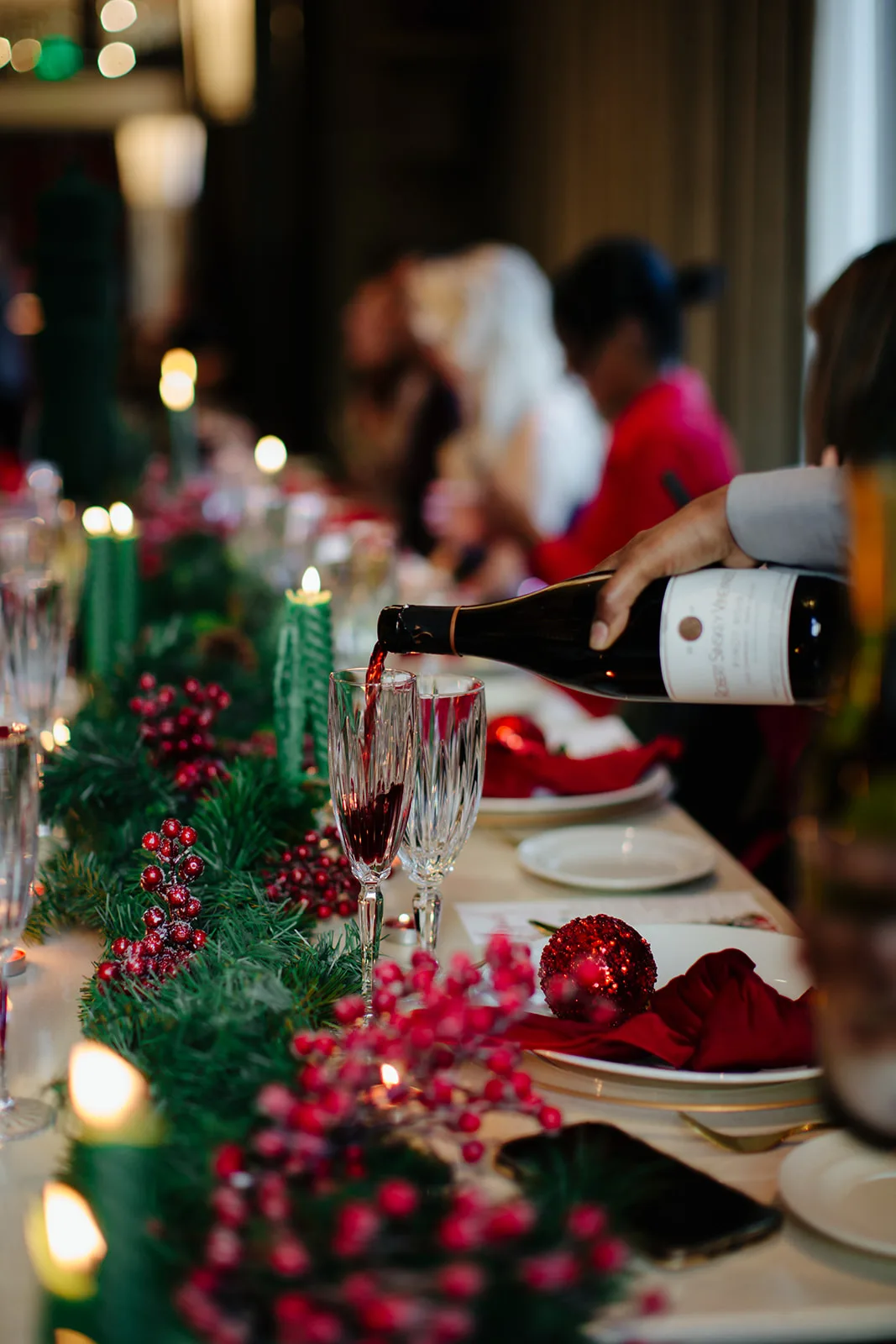 Red wine being poured into a crystal flute at a holiday dinner table with berries, garland and green candles