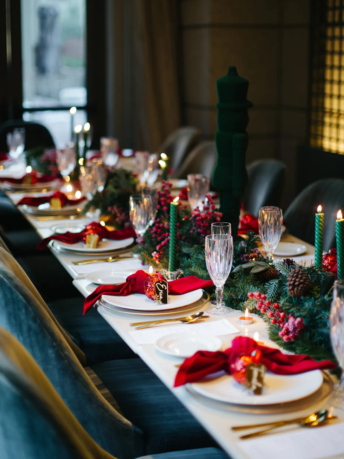 Holiday place setting with a red velvet napkin, glitter ornament name tag and gold flatware beside a pine and berry garland