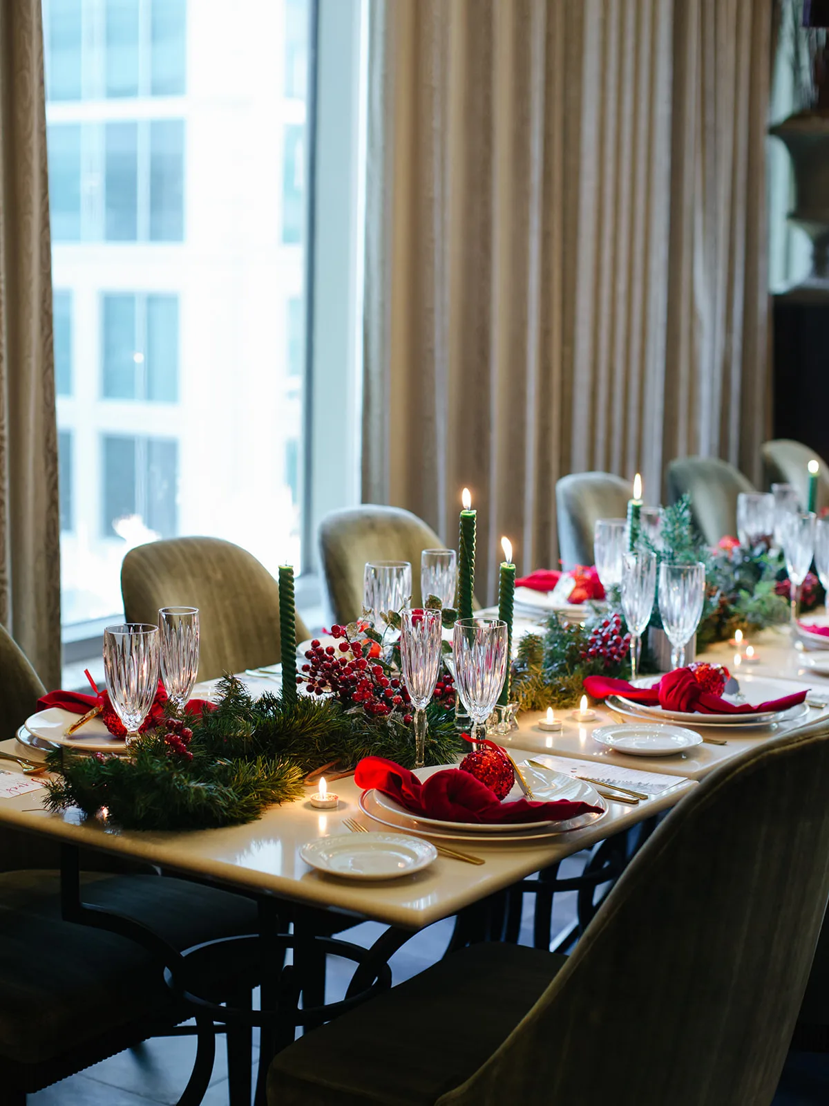 Candlelit holiday tablescape by a tall window with twisted green tapers, crystal flutes, berry garland and red velvet napkins