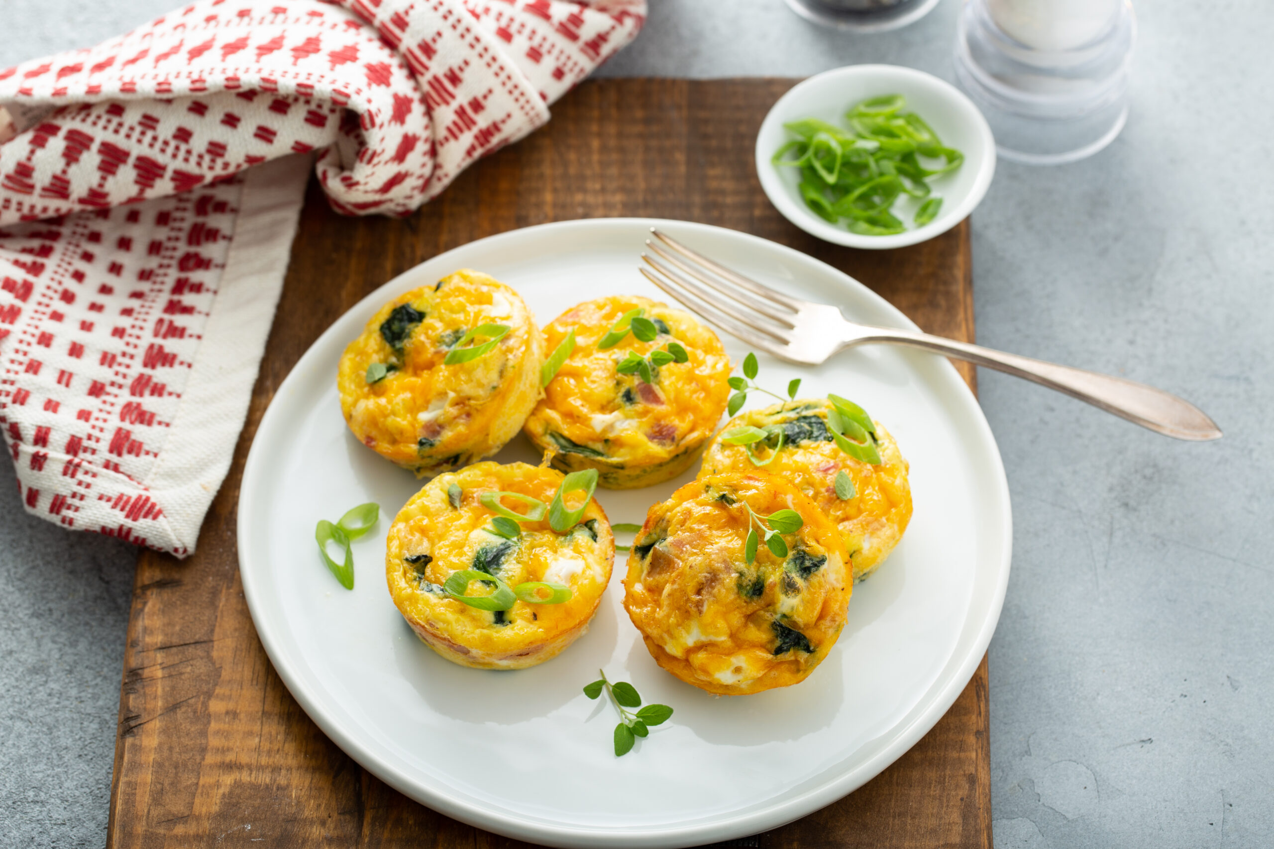 Four bacon, spinach and cheese egg muffins on a white plate with a fork and a bowl of sliced green onions