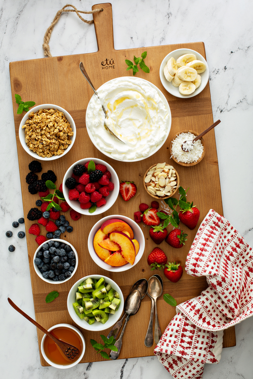 Greek yogurt board on a wooden board with bowls of yogurt, granola, berries, peaches, kiwi, coconut, almonds and honey