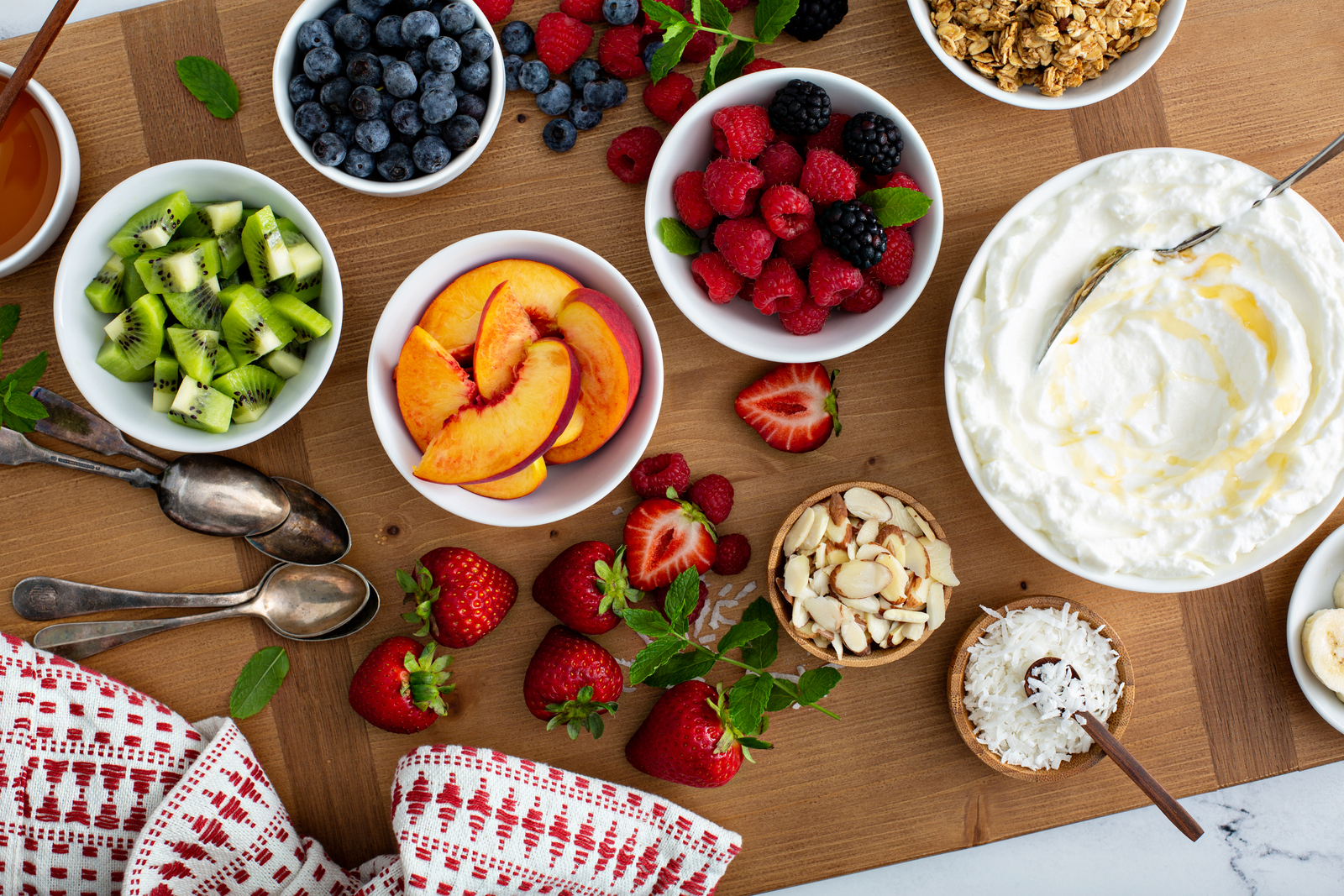 Overhead view of a yogurt board with bowls of blueberries, kiwi, peaches, raspberries, granola and whipped yogurt