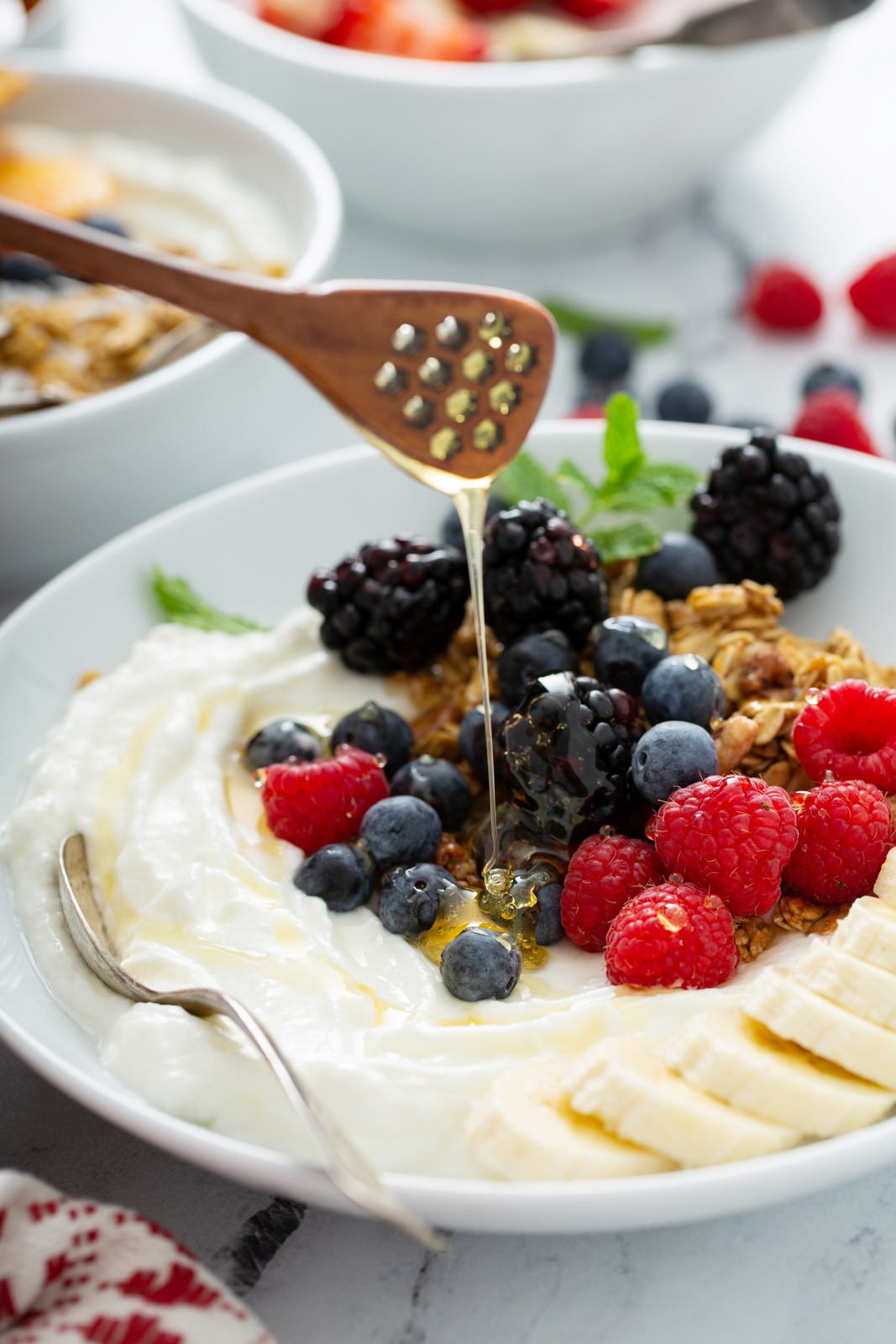 Honey being drizzled over a Greek yogurt bowl topped with blackberries, raspberries, blueberries, granola and banana